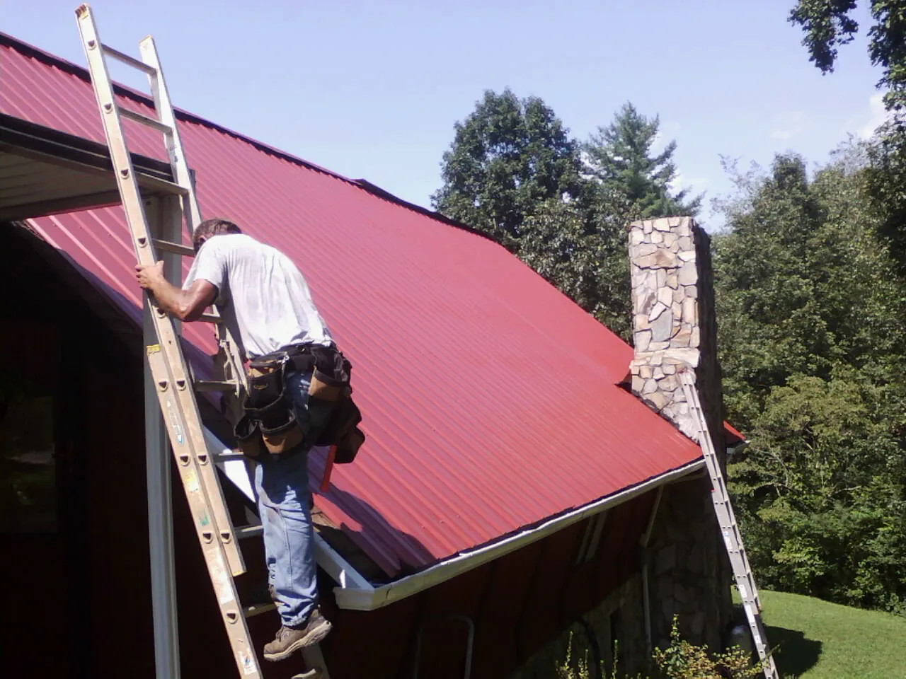 Roofer on a ladder beside a red metal roof and stone chimney — Soffit & Fascia Repair by Roofing Brothers in Robertsdale